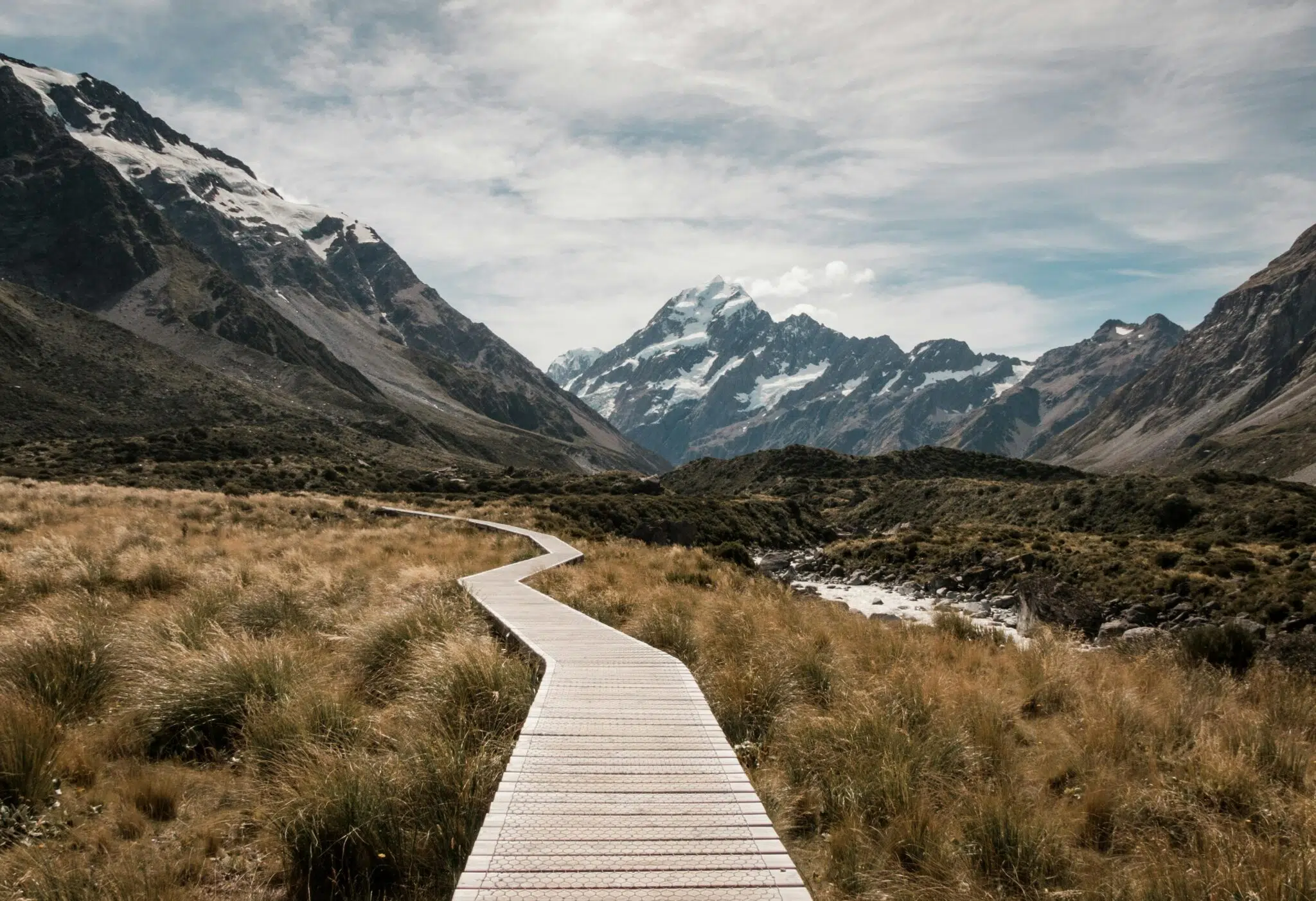 Brown Wooden Dock Path Surrounded With Green Grass Near Mountain Under White Clouds and Blue Sky at Daytime